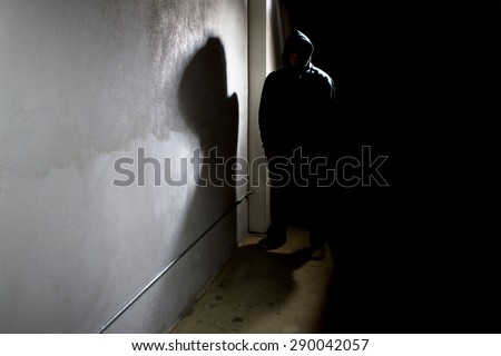 Silhouette of a hooded criminal stalking in the shadows of a dark street alley.  The man is a criminal waiting to ambush victims. The wall provides copyspace.