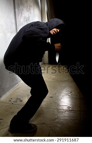 Hooded criminal with a knife hiding in the shadows of a street alley.  He is in a dark alley and partially visible as a silhouette.