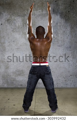 Fit black man in hip hop style clothing flexing back muscles. He is shirtless to show off his muscles and has a tattoo on one arm.