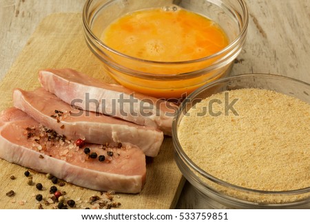 Three pieces of fresh raw pork chops, bowls with bread crumbs and eggs to the breading meat, peppercorns and salt crystals.Close,flat, horizontal view from above