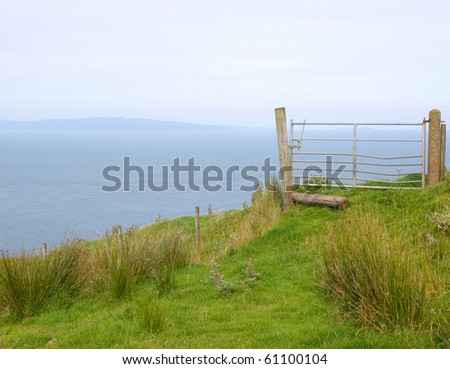 Field gate stands closed on the edge of a cliff overlooking the open sea with blue hills in the distance, illustrating the concept of a road to nowhere, end of the road, no entry, or danger.