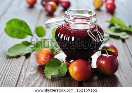 Plum jam in a glass jar and fresh fruits with leaves on wooden background closeup