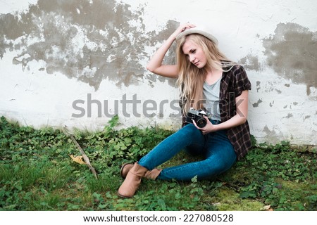 beautiful hipster girl in a hat with a vintage camera sits on a grass on a background of old wall
