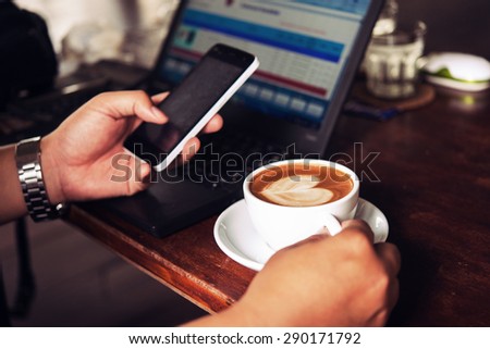 Businessman is working with laptop, holding a mobile phone in coffe shop and drinking coffe.