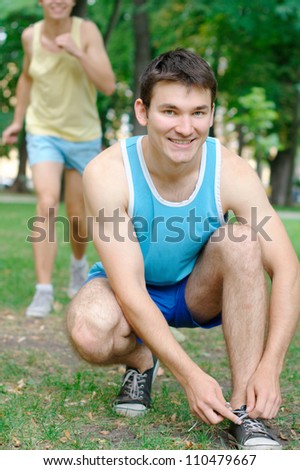 Young man doing shoelaces with another person jogging at the background at the park