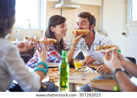 Two couples eating pizza in home, one girl is feeding guy with piece of pizza
