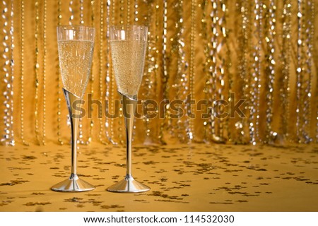 Two silver flutes in front of a shiny, sparkling, festive background hold bubbling champagne.