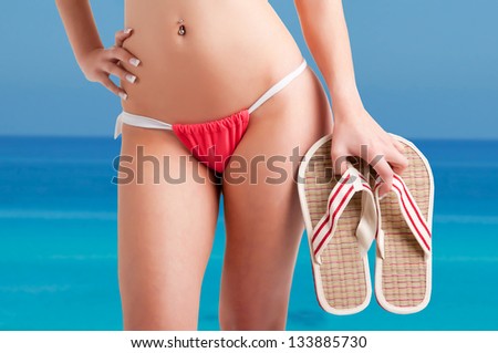 Woman wearing a bikini and holding flip-flops, with the ocean behind