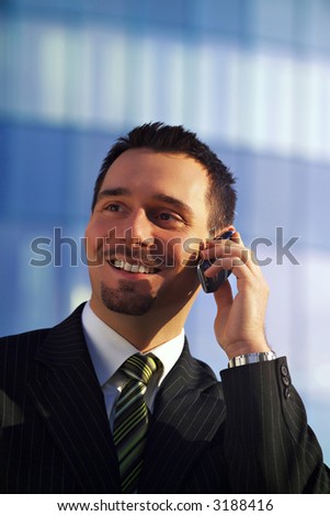 Attractive young businessman using a cell phone in front of a modern office building, smiling.