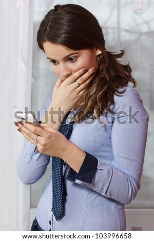 Portrait of a beautiful teenage girl with long brown hair in pony tail, looking at her mobile phone very surprised, indoors
