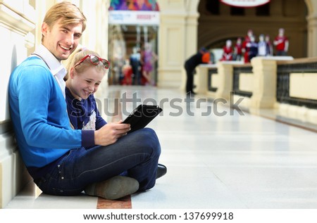 a young man and a little girl sitting on the floor of the mall and watch a movie on a digital tablet