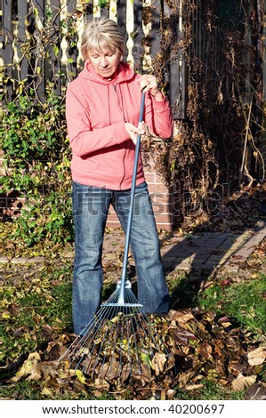 Senior woman raking autumn leaves in a lush garden.