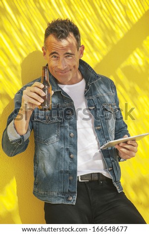 Smiling man drinking beer. / Senior man with digital tablet and beer on a yellow wall.