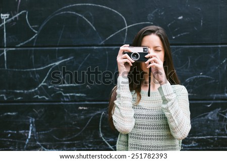 Beautiful woman standing in front a blackboard wall./ Young woman taking a photo with an old camera.