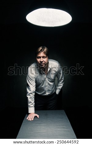 Man standing in dark room illuminated only by light from a lamp and looking in camera, hand on table