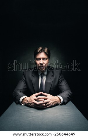 Man in suit sitting in dark room illuminated only by some light and looking in camera