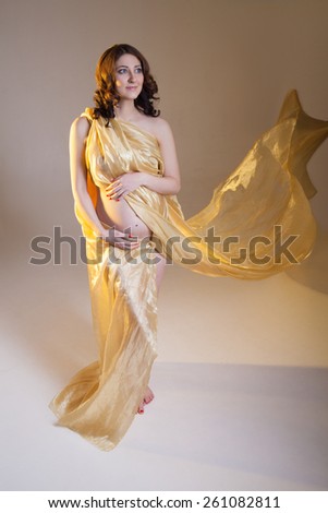 Beautiful young pregnant brunette woman with yellow transparent cloth in studio shot isolated on white background. Model is standing with her arms  on the belly.