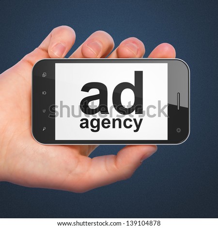 Marketing concept: hand holding smartphone with word Ad Agency on display. Generic mobile smart phone in hand on Dark Blue background.