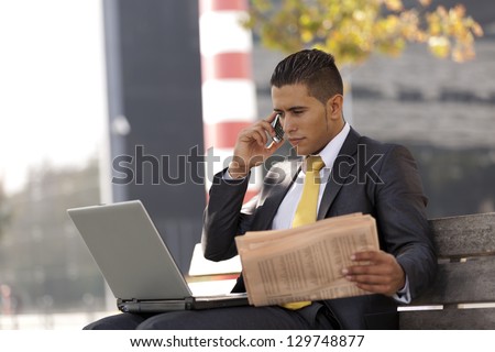 Businessman seated in a bench working with his laptop, calling at the cellular and reading his newspaper