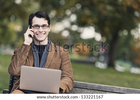 Young Man Sitting On Park Bench, Multi-tasking With Mobile Phone And Laptop Computer
