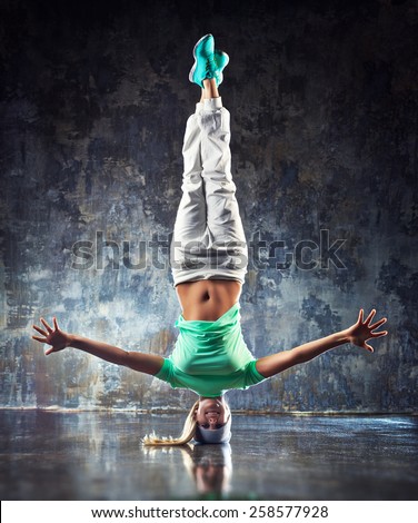 Young woman hip-hop dancer standing on head on wall background.