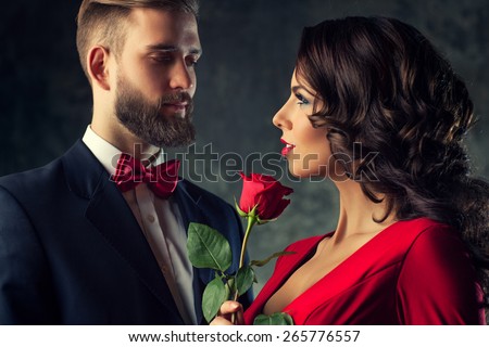 Young elegant couple in evening dress portrait. Woman in red holding rose and looking on man. Focus on woman.