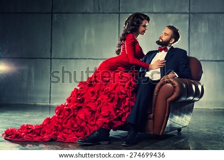 Young elegant loving couple in evening dress portrait. Woman in red and man in black suit sitting on chair.
