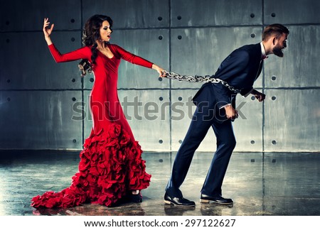 Young woman in red dress holding man on heavy chain. He tries to escape. Elegant evening clothing.