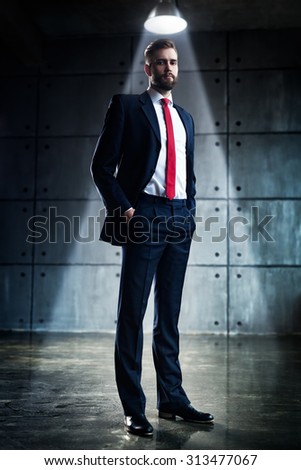 Young handsome businessman with beard in black suit standing under bright light in urban interior.