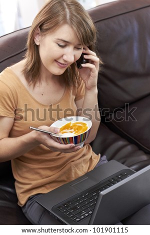 Happy young lady sitting on couch with laptop on her lap while eating healthy food and chatting on cellphone