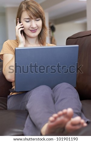 Barefoot lady sitting comfortably on couch while chatting on cellphone and working on laptop