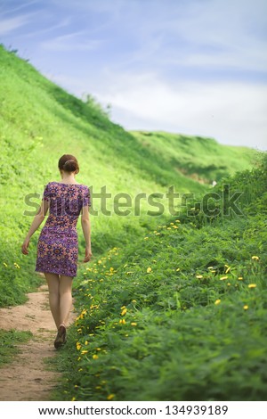 the girl goes on a footpath between hills covered with a fresh green grass. bright sunny day of summer.