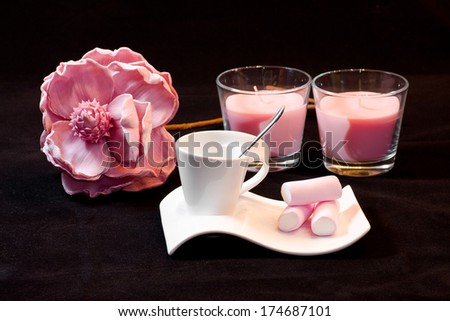 Composition with tea cup with marshmallows, pink rose and two pink candles over black background.
