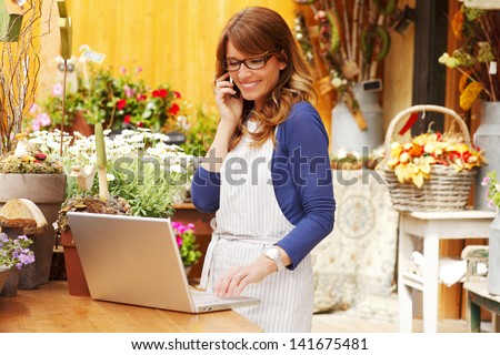 Smiling Mature Woman Florist Small Business Flower Shop Owner.  She is using her telephone and laptop to take orders for her store. Shallow Focus.