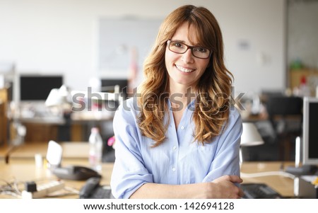 Smiling mature professional businesswoman in casual, with arms crossed standing in office. Shallow focus.