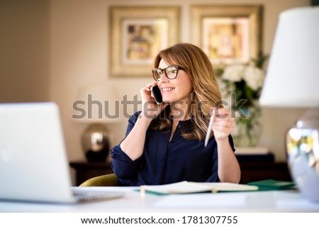 Shot happy businesswoman sitting at desk behind her laptop and talking with somebody on her mobile phone while working from home. Home office.