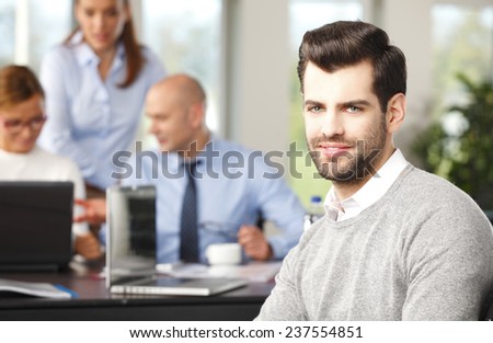 Portrait of successful young sales man sitting at meeting while business people working at background.