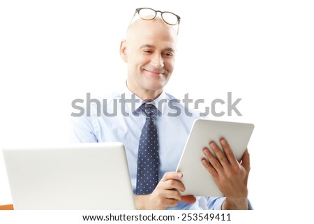 Busy sales man holding digital tablet while sitting at desk in front of computer. Isolated on white background.