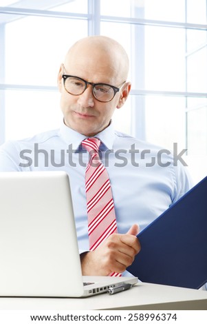 Portrait of busy businessman sitting at office in front of computer while holding file in his hand.