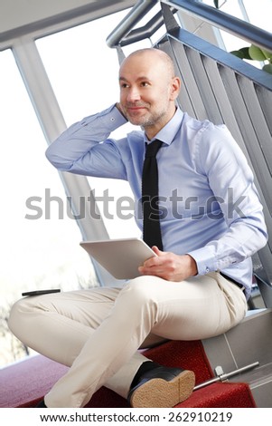 Portrait of executive businessman sitting at staircase at office while holding in hands digital tablet and relaxing.