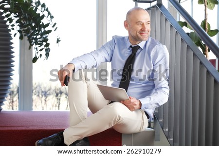 Portrait of executive businessman sitting at staircase at office while holding in hands digital tablet and relaxing.
