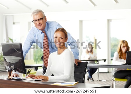Teamwork at office. Portrait of senior financial manager giving advise to businesswoman while sitting in front of computer and analyzing financial data.  At background sitting business persons.