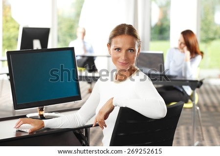 Business person working at office. Confident middle age businesswoman sitting at desk in front of computer and smiling at camera. Business people working at background.