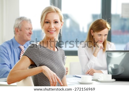 Portrait of attractive smiling businesswoman sitting at meeting while business people sitting at background and working on business report. Teamwork at office.