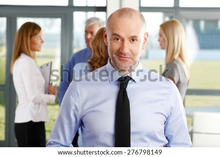 Close-up portrait of senior bank employee standing at office while business people discussing at background.