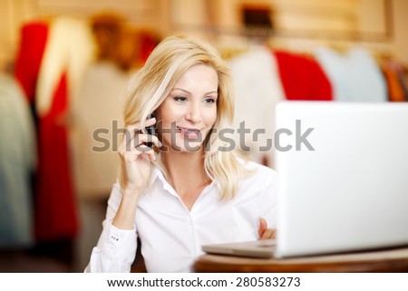 Small shop owner sitting in her store while working online at computer and making call.