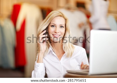 Portrait of small fashion store owner. Busy businesswoman sitting in her designer shop and making call while and using laptop.