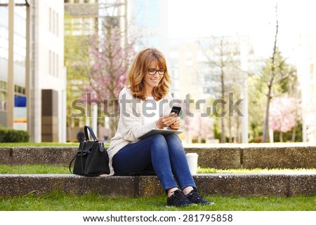 Businesswoman in front of an office building have a break.Typing message on smart phone and using her digital tablet.