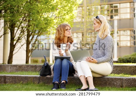 Image of two attractive sales woman sitting on the park and using laptop and digital tablet while talking at coffee break.