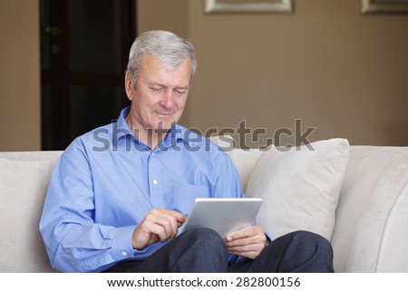 Portrait of senior man sitting at sofa at home with digital tablet and reading breaking news.